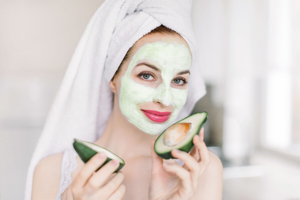 Woman holding avocado peaces, with a homemade avocado mask on her face and hair wrapped in bath towel