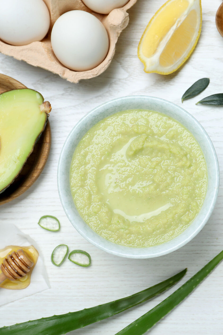 Homemade avocado face mask in bowl and ingredients on white wooden table